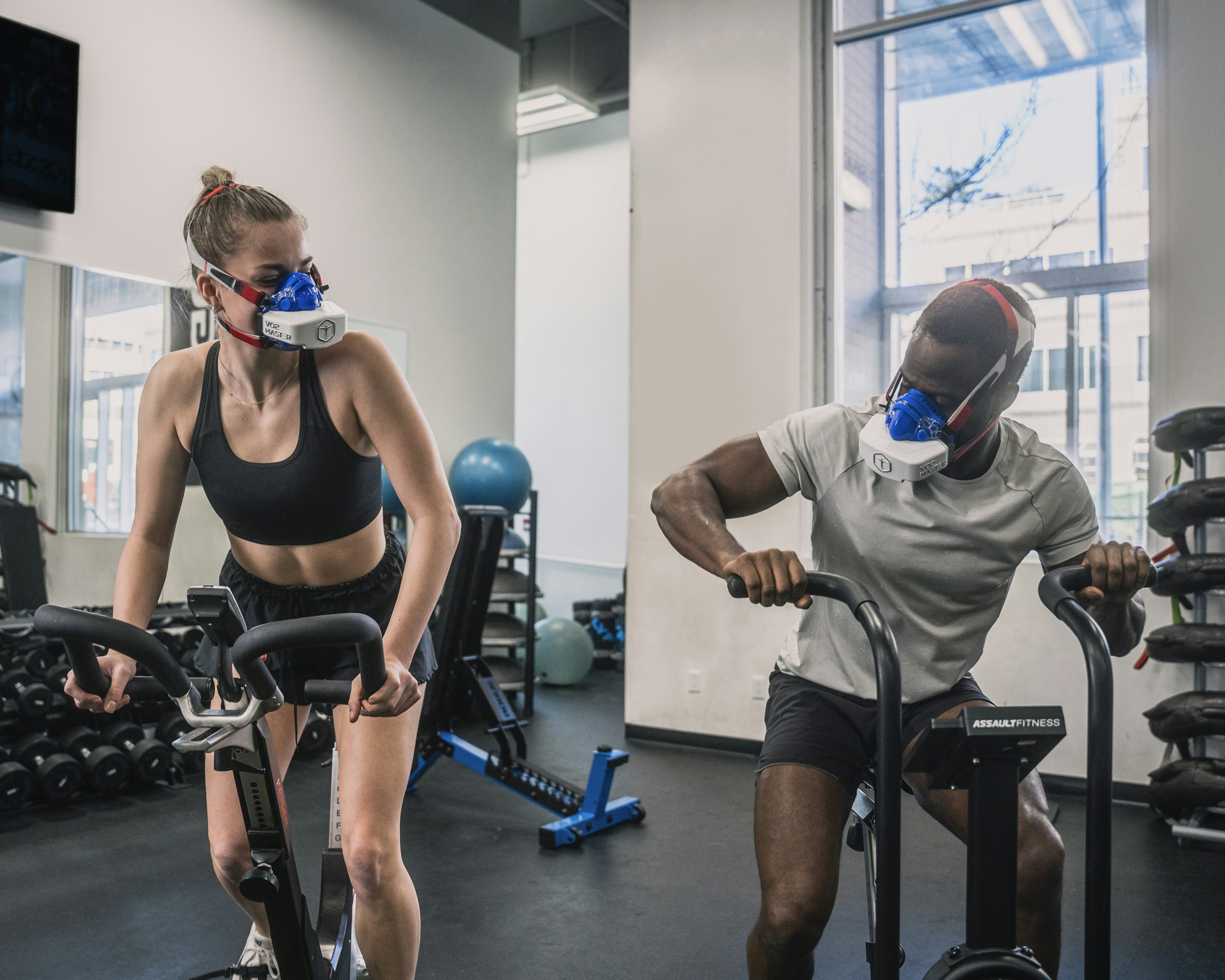 People side by side on exercise bikes, wearing CO2 measuring mask to evaluate cardio fitness. Peptides can improve sports performance